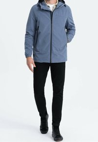 Giacca impermeabile azzurro chiaro con zip frontale, cappuccio regolabile, polsini elasticizzati e tasche laterali, abbinata a pantaloni neri slim-fit e sneakers.