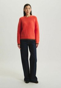 Pull en maille côtelée orange à manches longues et col rond. Associé à un jean flare bleu marine. Le pull présente des accents de bord en marron.