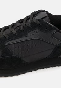 Zapatilla deportiva negra que combina gamuza y tejido texturizado, con detalles texturizados en el costado y una suela de goma robusta con ranuras.