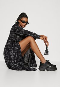 Zwarte jurk met stippen, lange mouwen en een gelaagde rok, gecombineerd met chunky zwarte schoenen. Kleine zwarte handtas als accent. Soepele stoftextuur.
