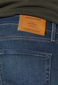 Mörkblå denimjeans med en läderetikett bak, med texten "JACK & JONES" och kontrasterande sömmar längs med skarvarna.