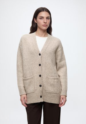 Femme portant un cardigan beige boutonné avec des poches avant sur un haut blanc et un pantalon foncé, debout devant un fond uni.