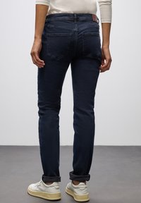 Tmavě modré, slim-fit džíny s hladkou texturou, s páskem v poloviční výšce a dvěma zadními kapsami. Otočené manžety a detaily z kůže.
