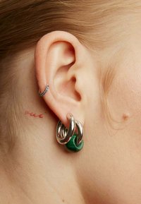 Pendientes de aro de metal plateado con un acento de resina verde, que presentan diseños entrelazados y una textura suave y pulida.