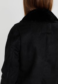 Czarny zamszowy płaszcz z puszystym czarnym kołnierzem. Charakteryzuje się dopasowanym krojem i teksturowanym materiałem, prezentującym bezszwowe szwy w okolicy ramion.