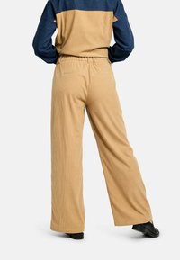 Személy bézs széles szárú kordbársony nadrágot és tengerészkék-bézs színblokkolt pulóvert visel, kezeit csípőjére téve, hátrafelé nézve.