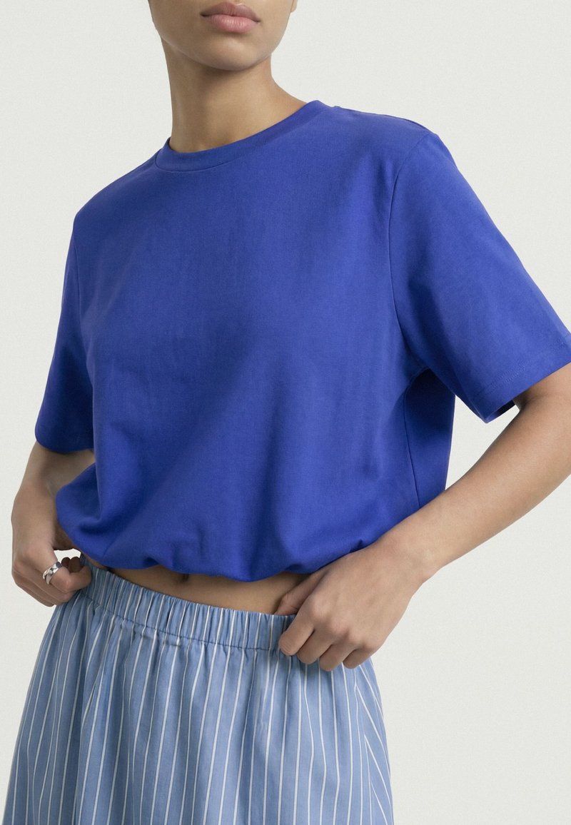 Personne portant une chemise à manches courtes bleu vif et un pantalon à taille élastique rayé bleu clair, les mains ajustant la taille.