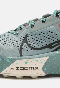 Close-up van een grijze en turquoise Nike ZoomX sneaker met gemusterde veters en gespetterde middenzooldetails.