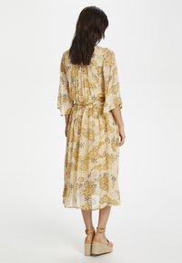 Abito maxi floreale in giallo chiaro con maniche a campana, vita elasticizzata, tessuto fluido e un motivo di fiori gialli e viola. Sandali con zeppa.