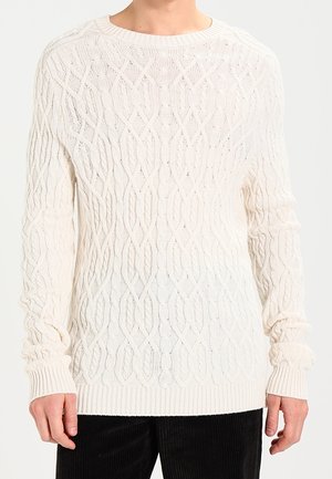 Mann trägt einen weißen Strickpullover mit Zopfmuster, langen Ärmeln und gerippten Bündchen sowie Saum, kombiniert mit schwarzen Cordhosen.