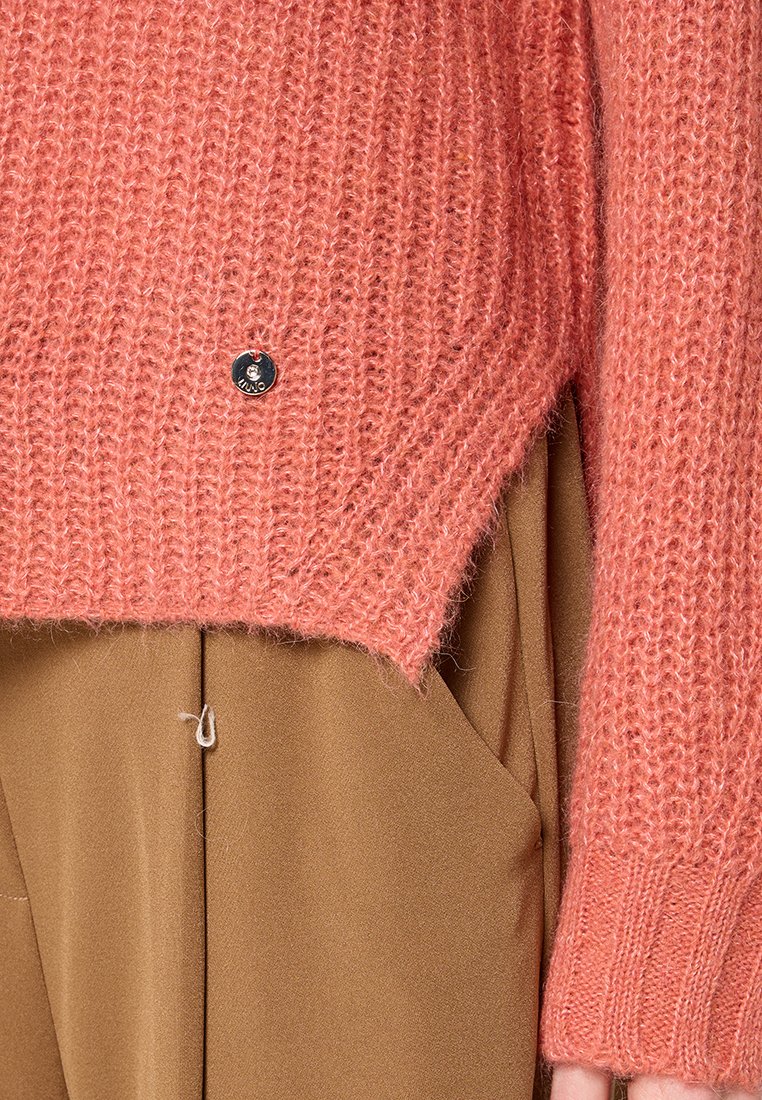 Maglione lavorato a maglia in soffice filato corallo con una texture a coste. Presenta un orlo leggermente più corto sul davanti e un accento in argento con il logo vicino alla cucitura.