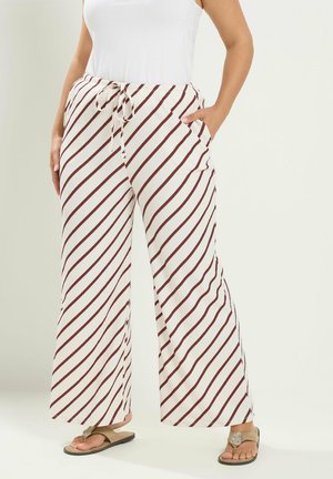 Frau trägt weiße, weit geschnittene Hose mit diagonalen braunen Streifen, kombiniert mit einem weißen ärmellosen Oberteil und beigen Sandalen.