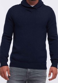 Pull en maille bleu marine avec col châle, poignets et ourlet côtelés. Présente une finition texturée. Associé à un jean gris clair.