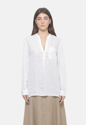 Donna con lunghi capelli castani indossa una camicia bianca a maniche lunghe con bottoni e una gonna beige, in piedi davanti a uno sfondo grigio chiaro.