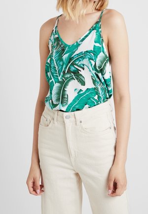 Vrouw met een mouwloze top met groene tropische bladprint en een hooggetailleerde off-white broek, staand tegen een effen achtergrond.