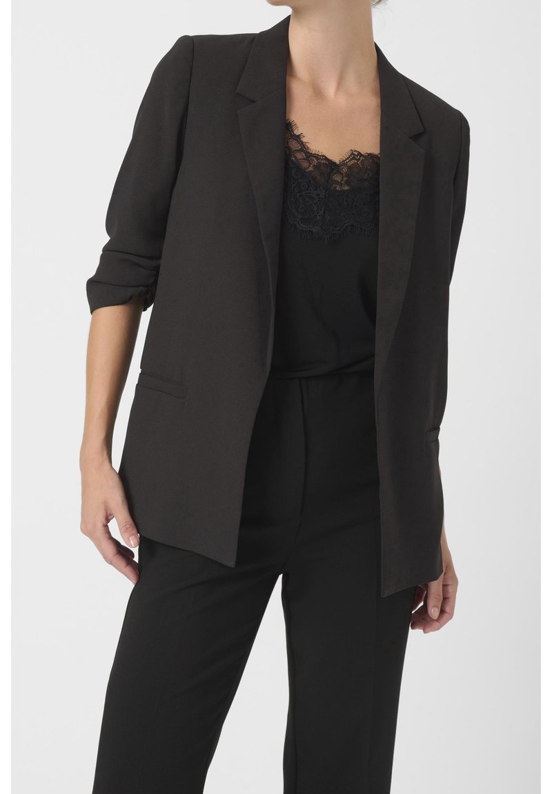 Black tailored blazer with a relaxed fit, notch lapels, rolled sleeves, and a single front pocket, paired with a black lace camisole underneath.