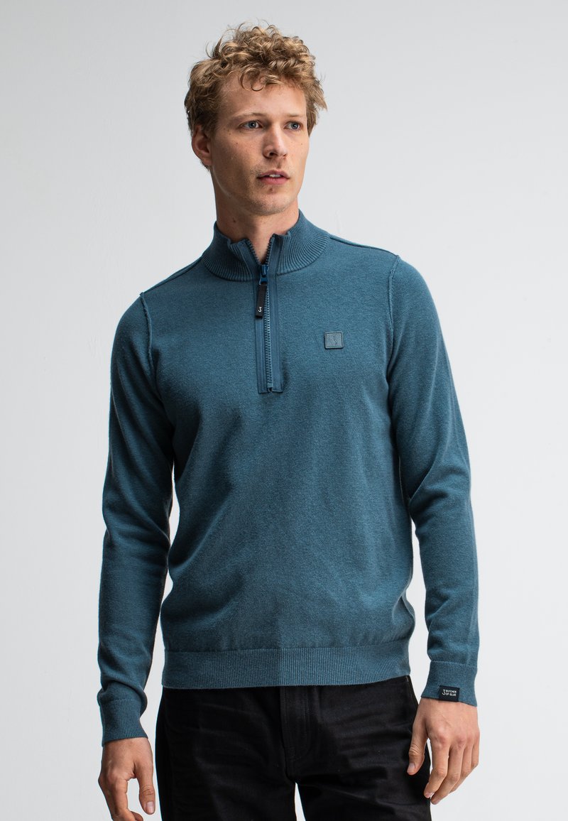 Türkiser Zip-Hals-Pullover aus einem weichen Material, mit einem gerippten Kragen und Bündchen sowie einem kleinen Logopatch auf der Brust. Sichtbare schwarze Hose.