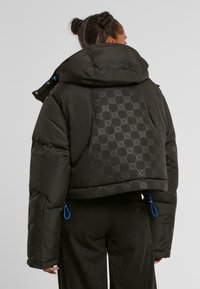 Sort puffjakke med oversized hætte, der har et tekstureret mønster på ryggen, blå hardware-detaljer og en cropped silhuet.