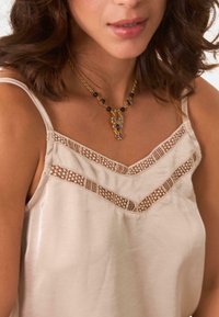 Camiseta sin mangas de crema con encaje en un patrón de espiga, con tirantes finos. Collier dorado con cuentas de colores y un colgante visible.