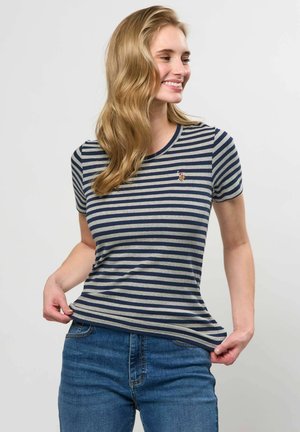 Femme aux longs cheveux blonds souriante, portant un t-shirt rayé bleu marine et gris ainsi qu'un jean bleu, tenant délicatement l'ourlet du t-shirt.