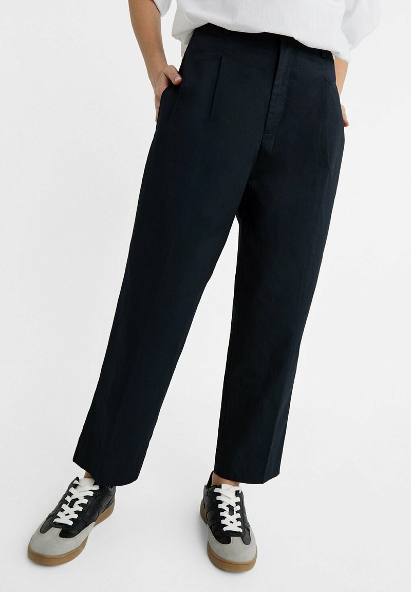 Pantaloni a gamba larga della marina con pieghe, realizzati in tessuto leggero. Indossati con sneakers nere e grigie con lacci bianchi.
