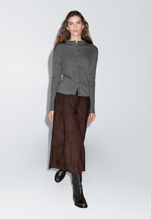 Cardigan en tricot gris avec des boutons, associé à une longue jupe marron et des bottes noirs hauteur genou. Texture lisse, design ajusté.