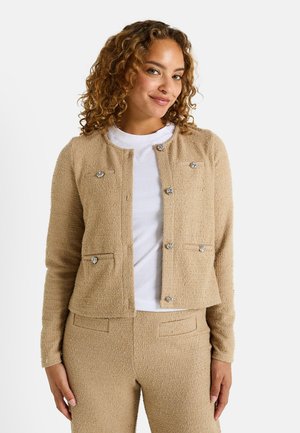 Cárdigan texturizado beige con cuello redondo, bolsillos frontales y acentos de botones plateados. Llevado sobre una camisa blanca, con un diseño recortado.