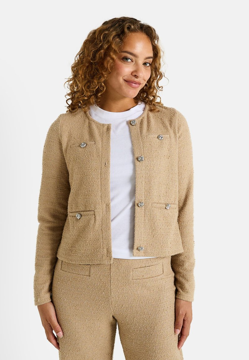 Cárdigan texturizado beige con cuello redondo, bolsillos frontales y acentos de botones plateados. Llevado sobre una camisa blanca, con un diseño recortado.