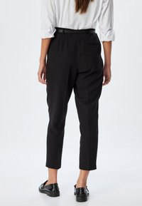 Pantaloni neri con una vestibilità dritta, caratterizzati da una vita media, passanti per la cintura e due tasche posteriori, abbinati a scarpe nere lucide.