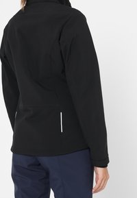 Veste softshell noire avec un col haut, texture lisse et léger éclat. Comprend deux poches latérales avec des zips blancs.