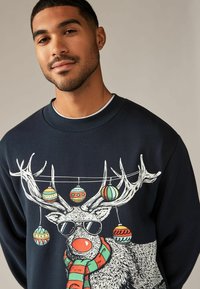 Felpa blu navy con grafica di un rinoceronte dotato di occhiali da sole, decorata con ornamenti colorati e una sciarpa a righe.