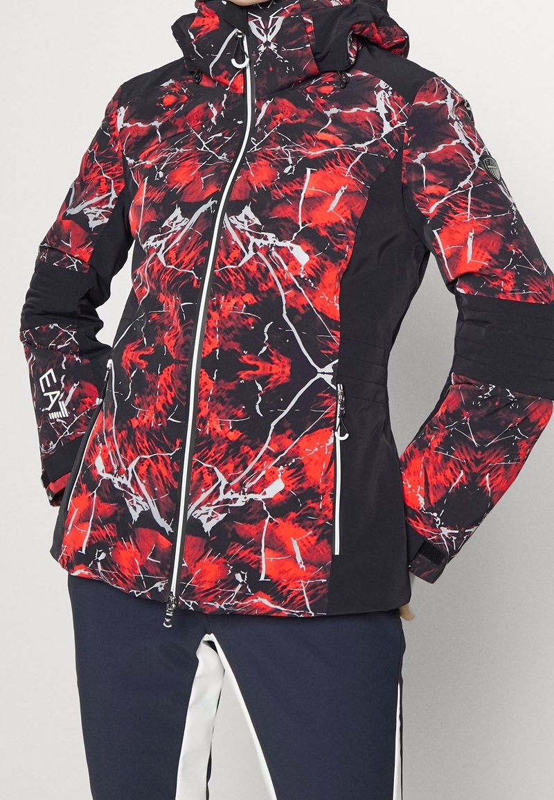 Huva jacka med svart bakgrund, med ett livligt röd och vit abstrakt mönster, med dragkedja fram och randiga detaljer på ärmarna.