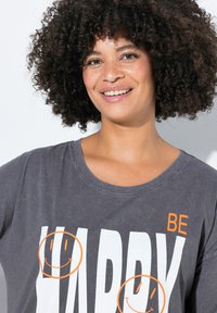 Grijze t-shirt met witte bedrukte tekst "HAPPY" en oranje glimlachende gezichten, voorzien van een ronde halslijn en korte mouwen, met een gladde stoftextuur.