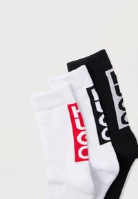 Tri páry žíhaných ponožiek, dve biele s červenými a čiernymi logami, jedna čierna s bielym logom, usporiadané prekrývajúc sa na svetlom pozadí.