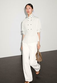 Lys grå, stribet bluse med høj krave og sorte knapper, parret med løse hvide bukser og en brun ruskinds taske. Glatte teksturer, minimalistisk design.