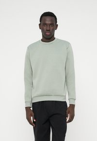 Mladý muž stojí čelom dopredu, oblečený v svetlo šedozelenom mikinách a čiernych nohaviciach na bielom pozadí.