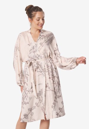 Kobieta w jasnoróżowej kwiatowej sukience kopertowej z długimi rękawami i wiązaniem w talii, uśmiechnięta i pozująca na jednolitym tle.
