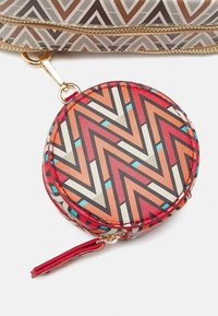 Compatto rotondo con motivo a zigzag multicolore con accenti rossi. Materiale sintetico liscio. Dotato di cerniera e tracolla rossa per la portabilità.