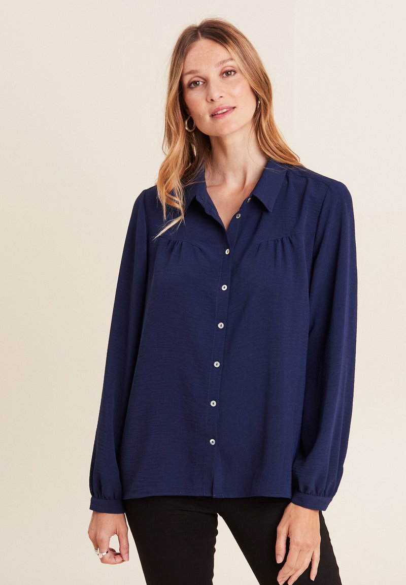 Camicia abbottonata blu navy con maniche lunghe, dotata di colletto, pieghe sul petto e una texture liscia. Semplici bottoni bianchi decorano la parte frontale.