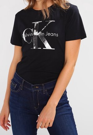 Kobieta w czarnej koszulce Calvin Klein Jeans z dużym logo "CK" oraz ciemnoniebieskich dżinsach na jasnym, jednolitym tle.
