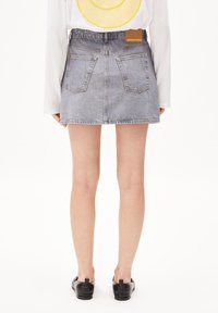 Jupe en denim bleu clair, avec une taille, deux poches arrière et une finition texturée. Portée avec une chemise blanche à manches longues et des chaussures noires.