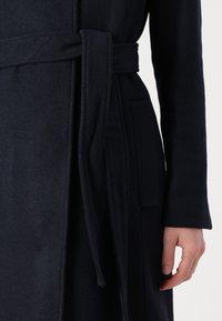 Lång mörk marinblå kappa i mjuk ullblandning, med ett knytbart bälte, sidofickor och en slät yta, delvis visad med en hand som håller i bältet.