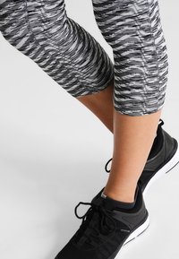 Kolečkově vzorované černobílé legíny s hladkou texturou, které jsou spárovány s černými sportovními botami s bílou podrážkou a tkaničkami.