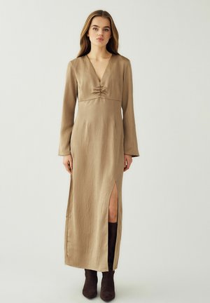 Vrouw draagt een lange beige jurk met een split aan de voorkant en lange mouwen, gecombineerd met donkerbruine laarzen, staand tegen een effen achtergrond.