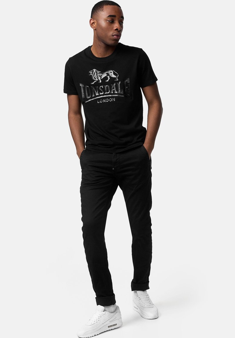 Camiseta negra de manga corta con un logo de león plateado y el texto "Lonsdale London". Se lleva con pantalones negros y zapatillas blancas.