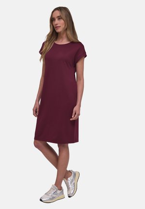 Kvinde iført en knælange, kortærmet vinrød kjole kombineret med sølvfarvede sneakers, stående med det ene ben let krydset.