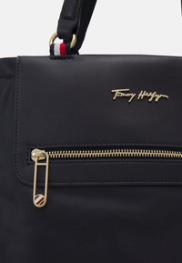 Borsa a tracolla in nylon nero con una cerniera dorata, un logo frontale e una tracolla decorativa tricolore. Superficie texturizzata con accenti lisci.