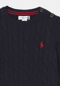 Granatowy sweter z dzianiny z wzorem w warkocze, z dwoma dekoracyjnymi guzikami na ramieniu oraz małym czerwonym logo na piersi.