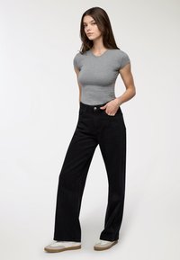 T-shirt grigio aderente, jeans neri a vita alta e sneakers bianche con suola marrone. La modella è in piedi con una mano sui fianchi su uno sfondo chiaro.