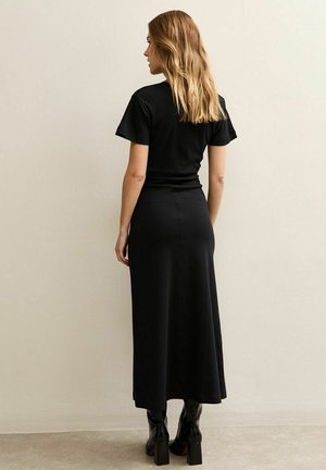 Vrouw met lang blond haar, gekleed in een zwarte nauwsluitende top met korte mouwen en een bijpassende lange rok, met zwarte laarzen met hakken, staand tegen een effen muur.